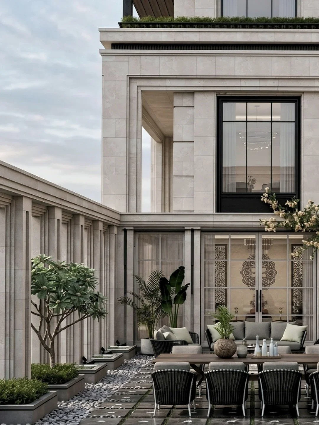 Exterior view of a modern luxury residence with a patio featuring plants, a dining area with a wooden table, black chairs, and beige cushions. Large windows and architectural details are visible on the building facade.