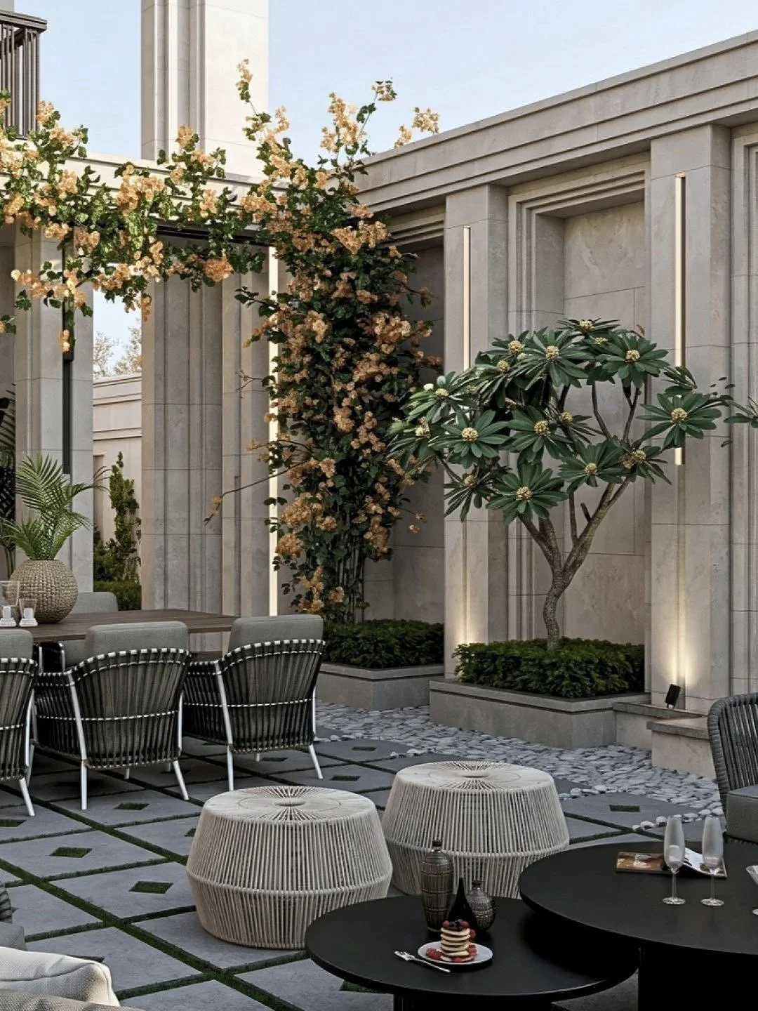 Outdoor patio with modern furniture, including black round tables, patterned seating, and decorative planters, featuring a flowering vine and a tree against a light stone building wall.