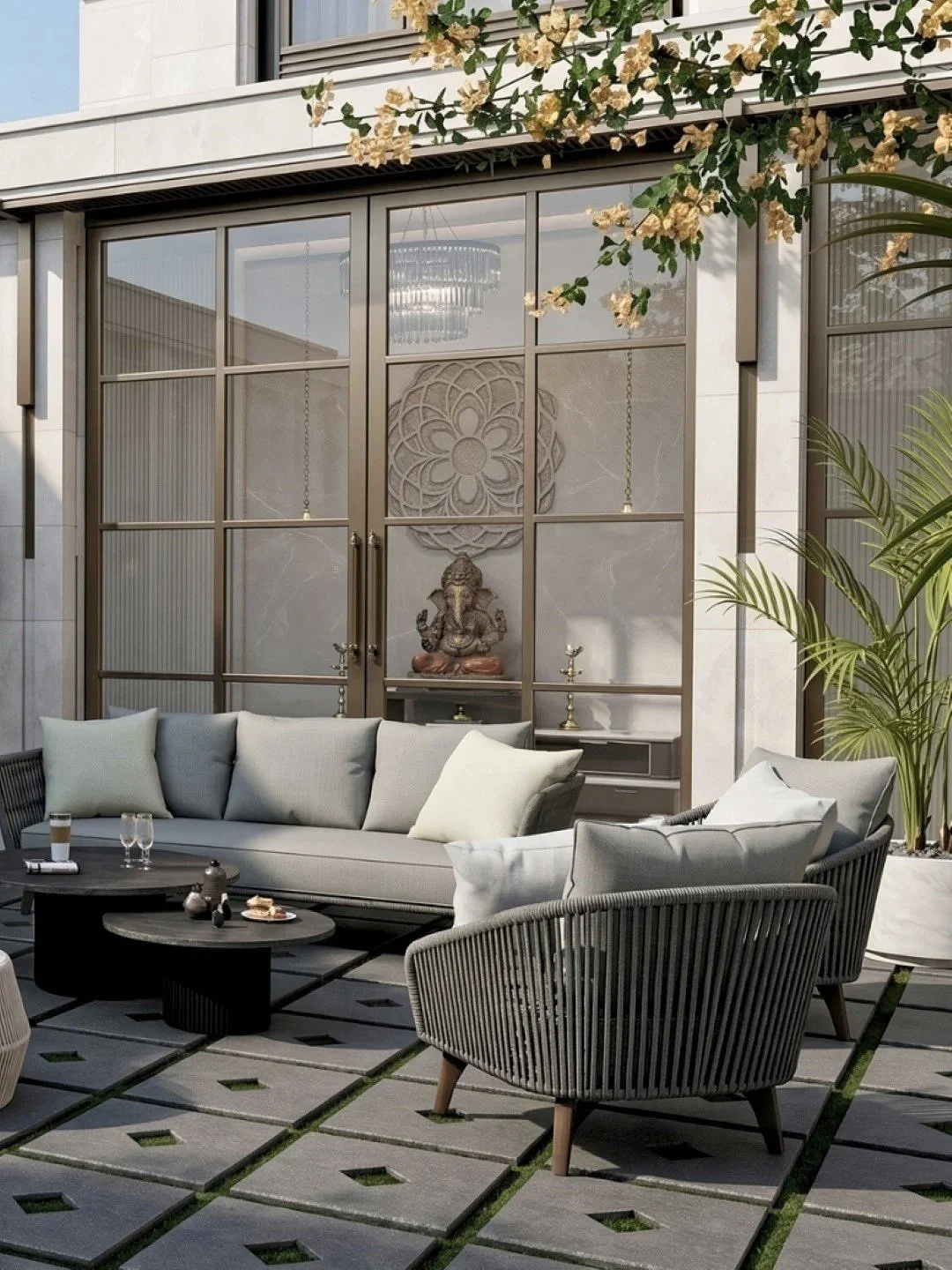 Outdoor patio with modern grey sofa and chairs, black coffee tables, decorative pillows, a Buddha statue, hanging candle holders, potted plant, and a glass sliding door with a floral design, greenery, and flowers above.