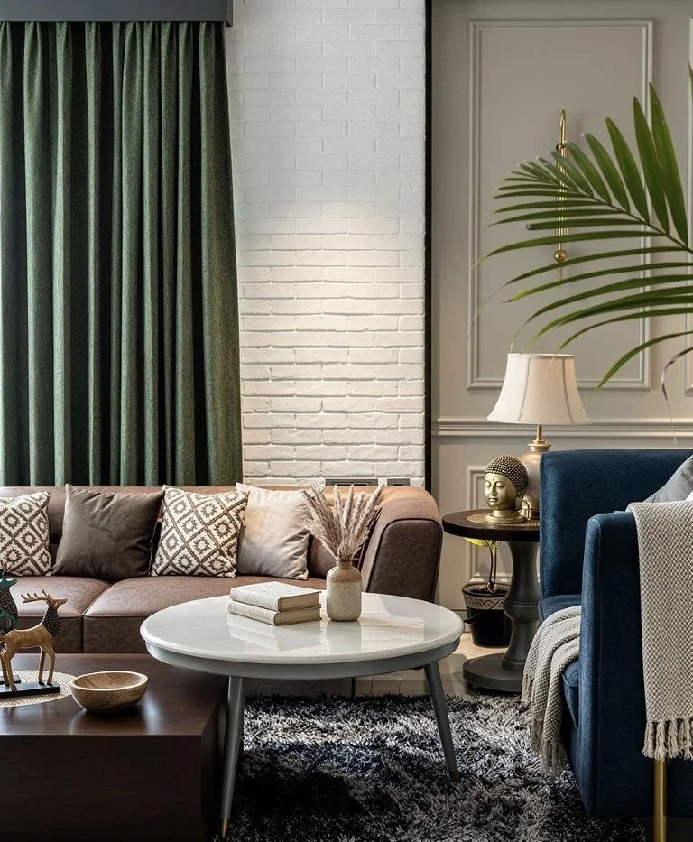 A cozy living room with a brown sofa, patterned pillows, a white round coffee table with books and a decorative vase, a side table with a Buddha head sculpture and lamp, a beige wall with molding, green curtains, and a blue armchair with a blanket, a