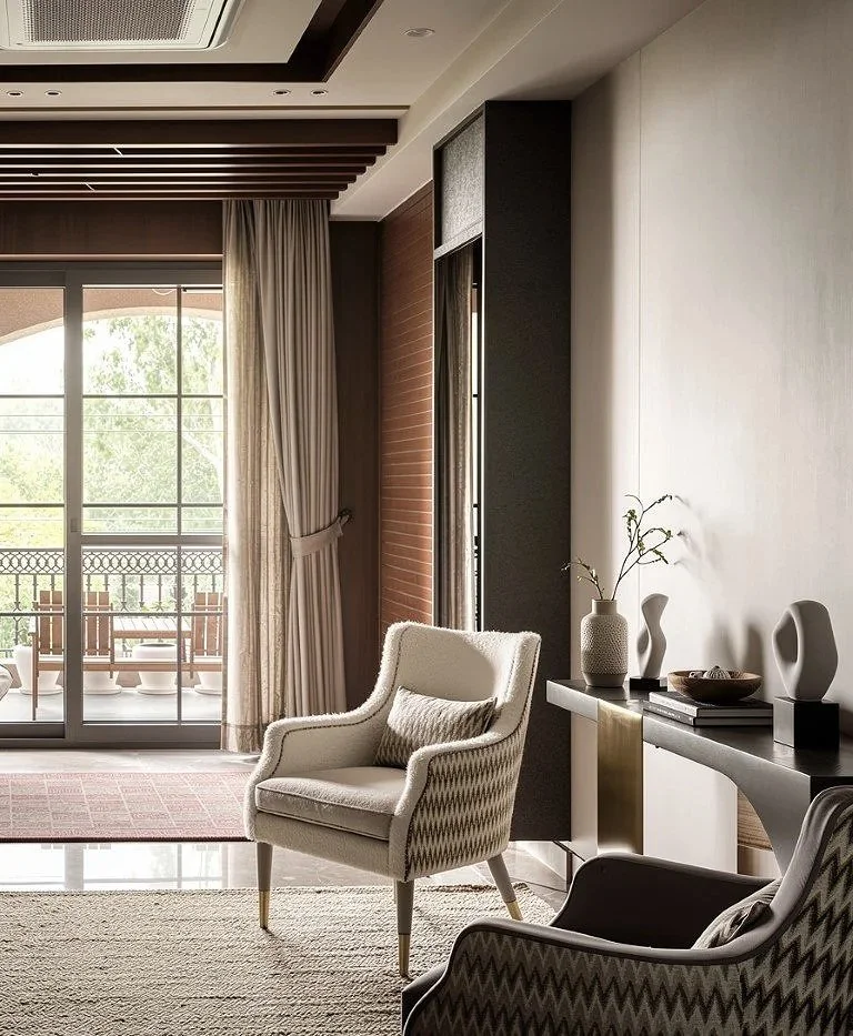 A living room with beige armchairs, decorative vases, a wooden bowl, and a side table, with large glass doors opening to a balcony with outdoor seating and greenery outside.