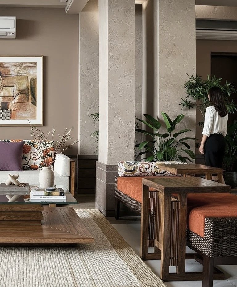Modern living room with a sofa, coffee table, decorative pillows, empty and patterned ottomans, a woman standing near the doorway, artwork on the wall, and an air conditioning unit.