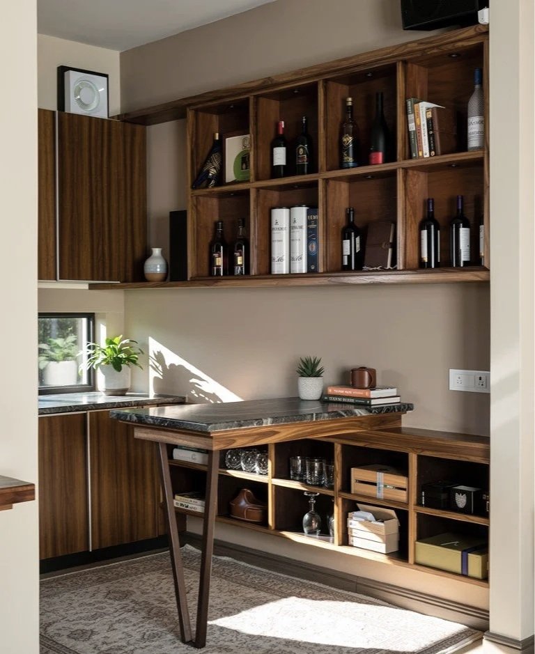 Wooden bar or wine cabinet with bottles of wine and liquor, some books, decorative items, and a small potted plant, situated in a cozy room with a window and a rug.