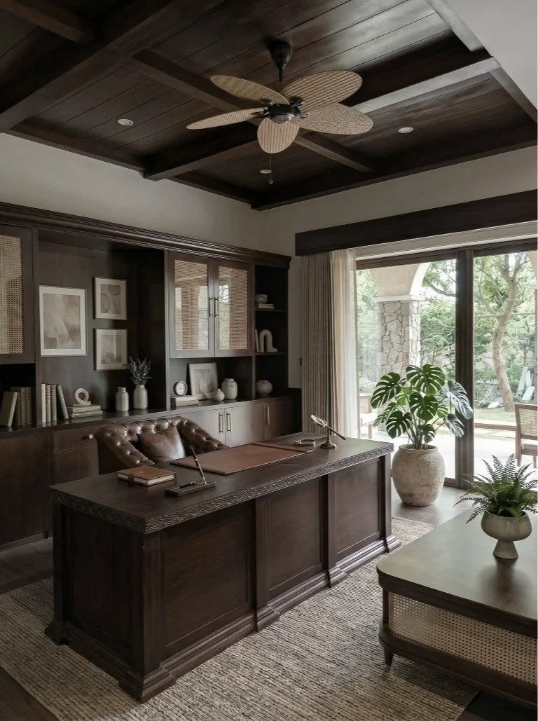 A home office with a dark wood desk, brown leather tufted chair, a large potted plant near sliding glass doors, built-in dark wood shelves with decorative items, and a ceiling fan with wicker blades.