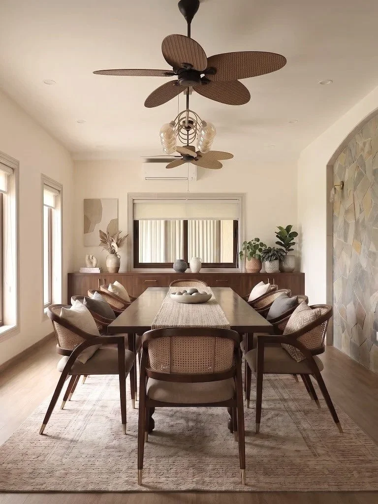 A modern dining room with a long wooden table, eight upholstered chairs, decorative vases, and plants against a neutral background with art on the walls and large windows.