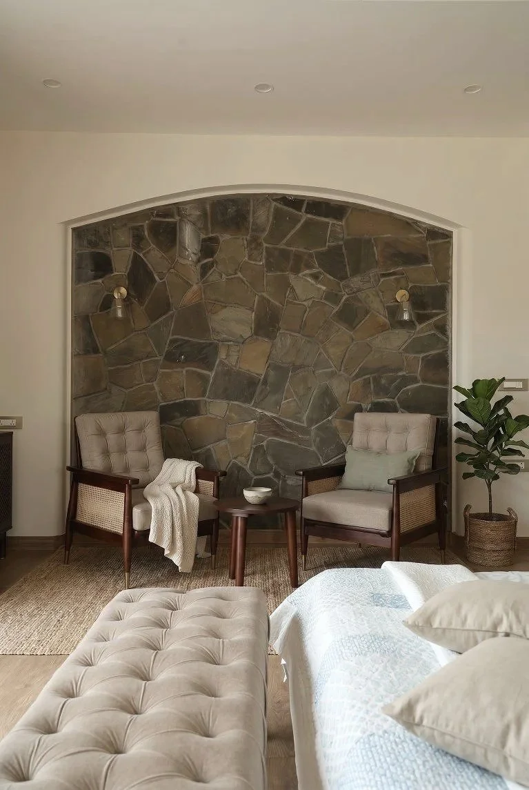 A cozy living room corner featuring two beige upholstered armchairs with wooden frames, a small round wooden side table between them, a potted plant in a woven basket, and a stone accent wall with two wall-mounted light fixtures. Part of a bed and a 