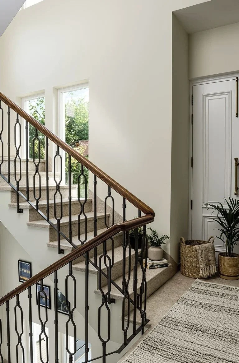 Interior view of a staircase with a wooden handrail, beige carpet, large windows, and decorative plants near a white door.