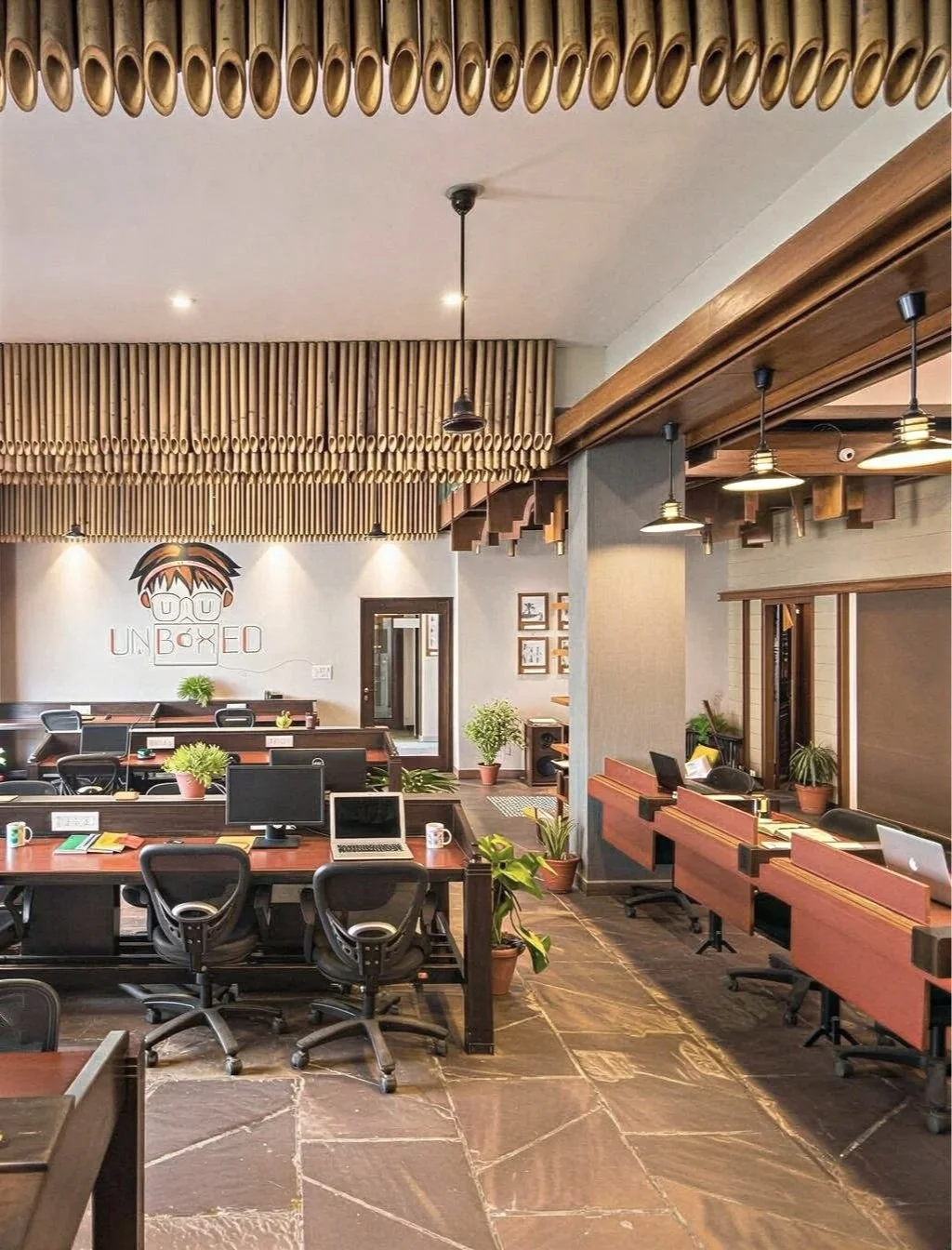 Modern office with work desks, chairs, laptops, and plants, featuring wooden ceiling accents and a logo on the wall.