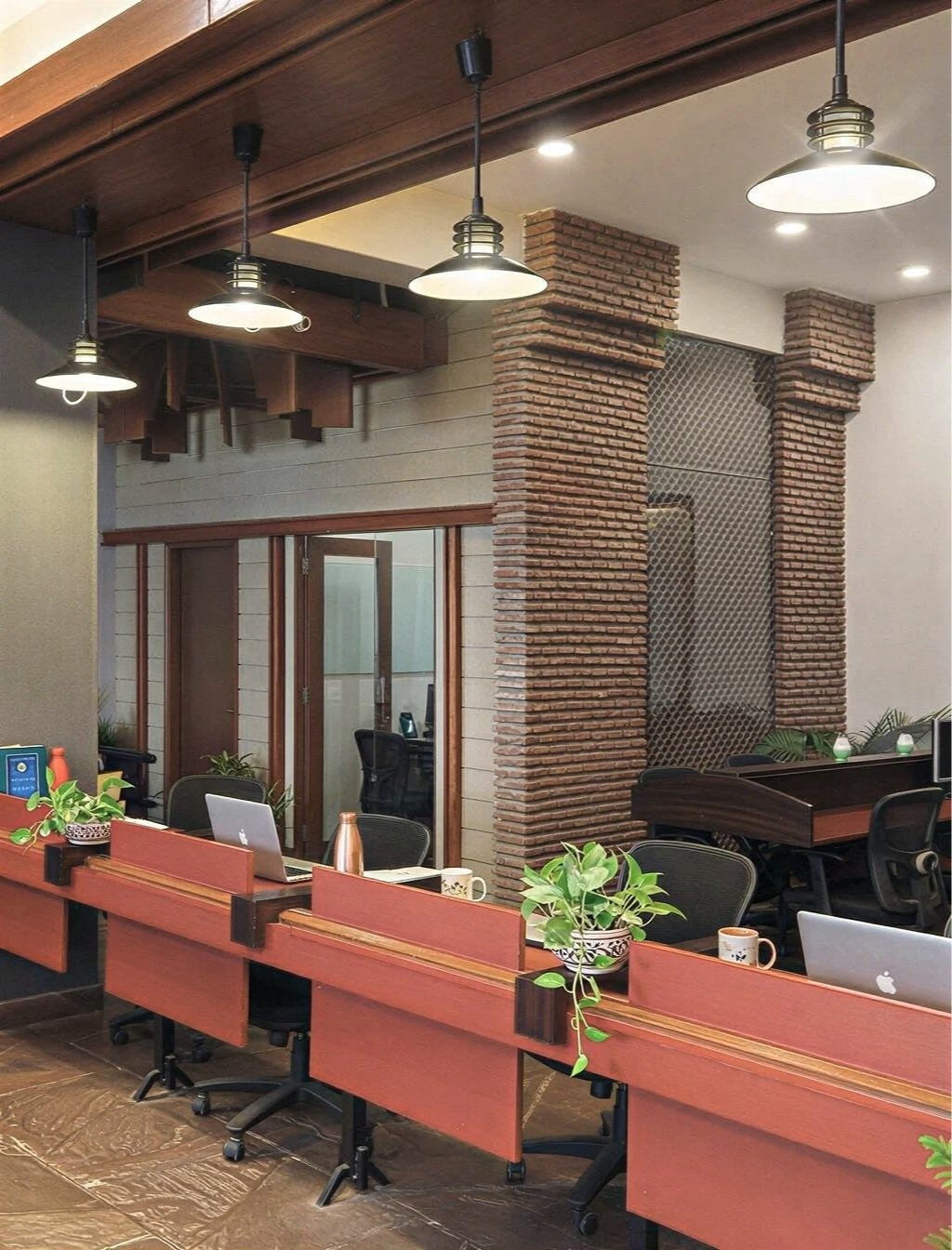 Modern office space with work desks, chairs, laptops, potted plants, and hanging pendant lights. The decor includes exposed brick columns and a wooden ceiling.