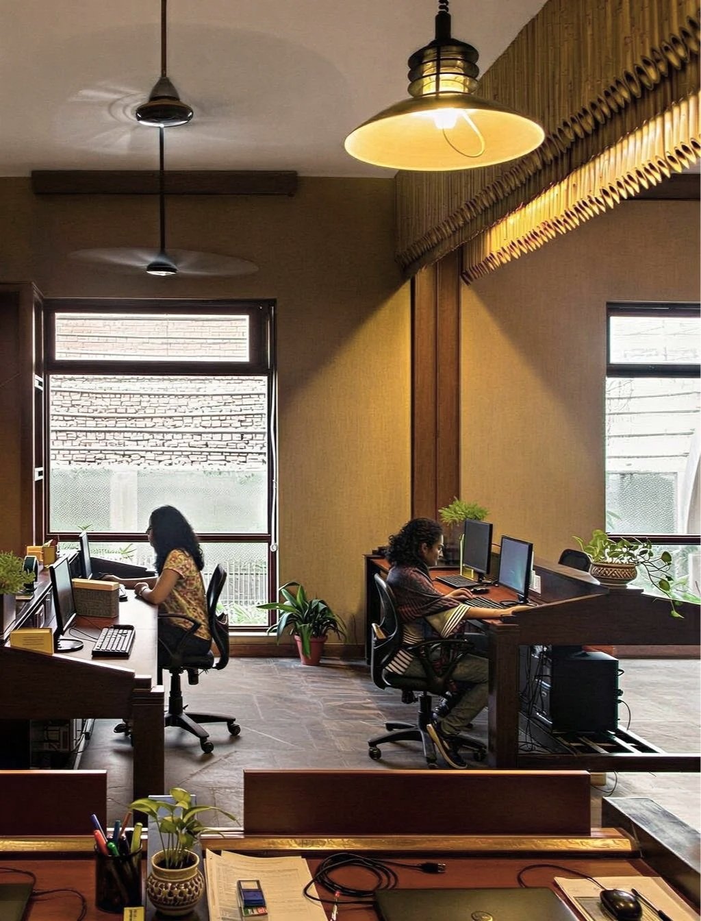 Two women working at desks with desktop computers in a warmly lit office with large windows and potted plants.
