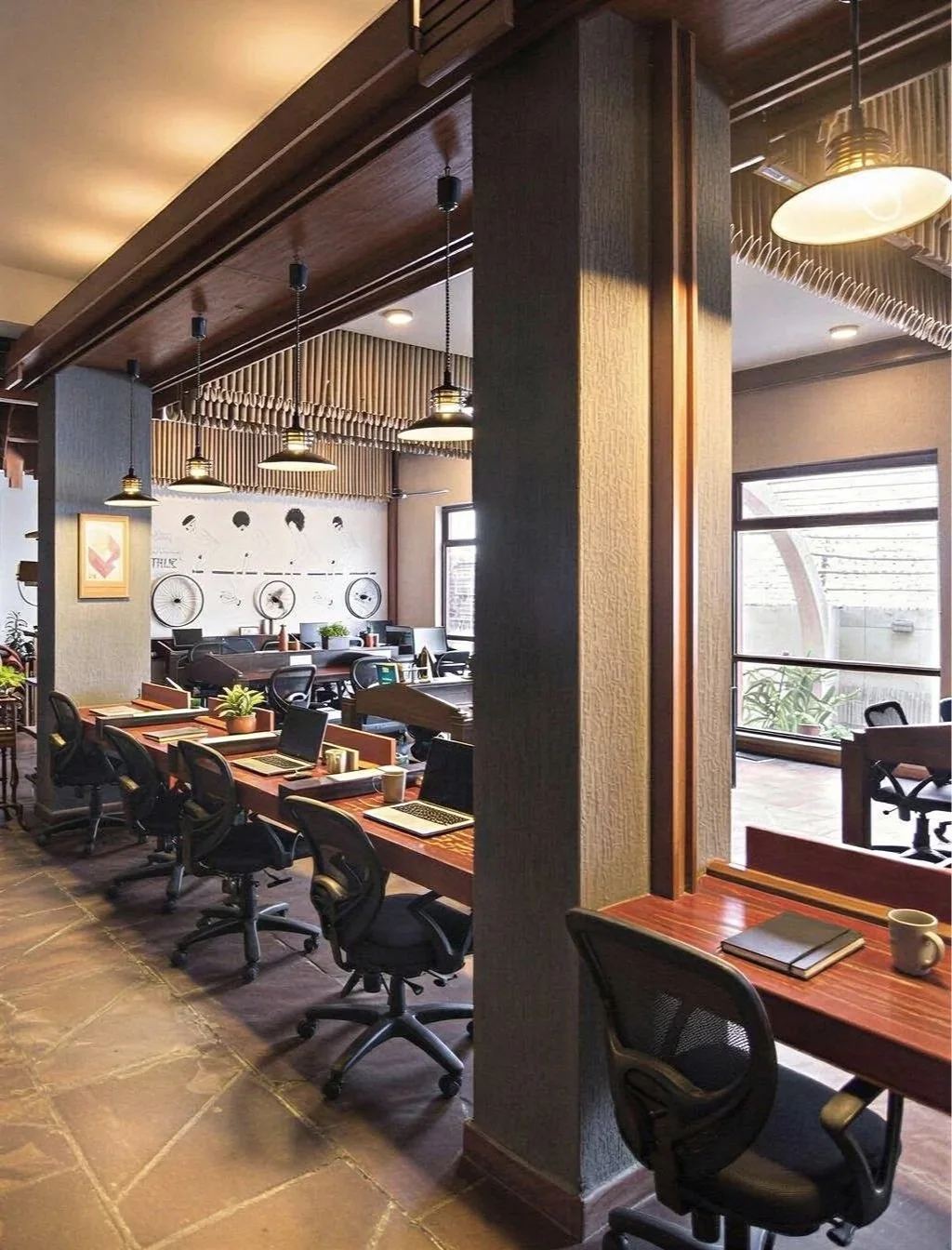 Interior of a modern office or co-working space with a large conference table, multiple chairs, computers, notebooks, and a cup on a small wooden desk. There are pendant lights, a wall with artistic decoration, and large windows letting in natural li