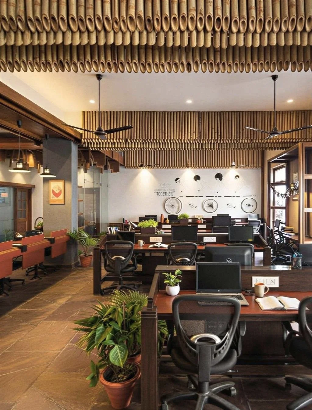 Modern office space with multiple desks and computer monitors, black office chairs, potted plants, wooden ceiling and wall decor, and wall art featuring bicycles.