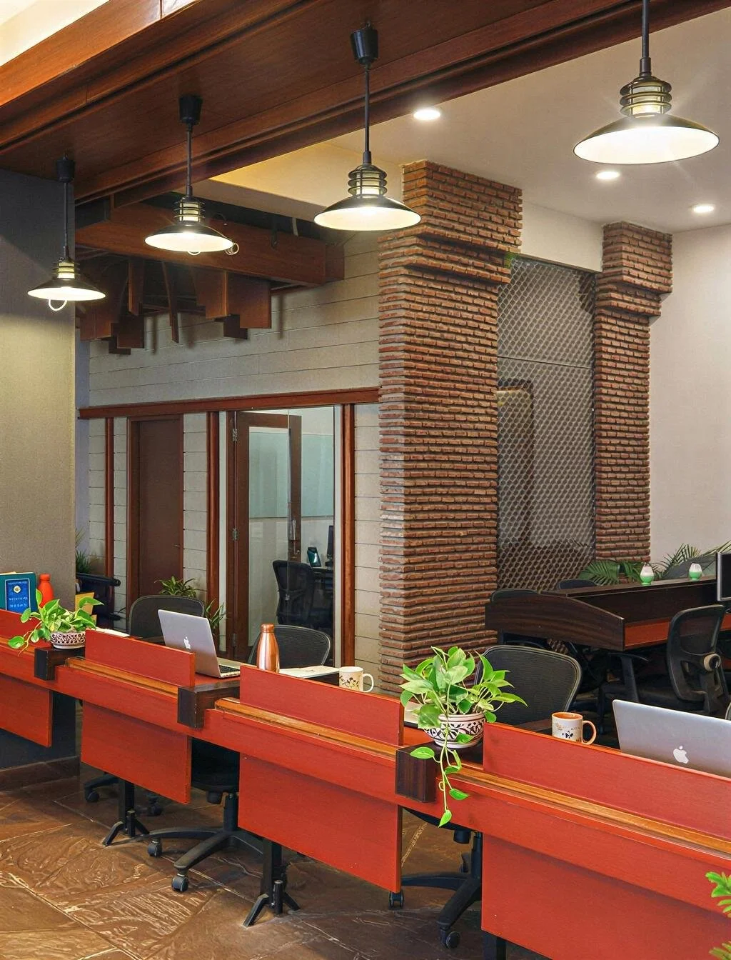 Modern office space with work desks, chairs, laptops, potted plants, and hanging pendant lights. The decor includes exposed brick columns and a wooden ceiling.