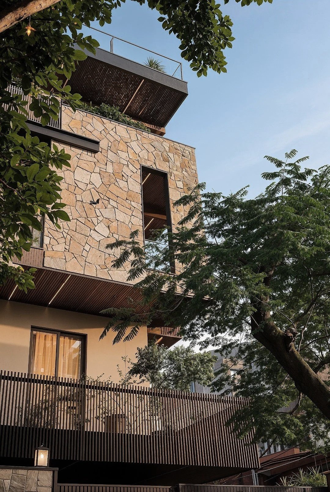 A modern multi-story building with a stone facade, large windows, and wooden accents, surrounded by trees with green leaves. The sky in the background is clear and blue.