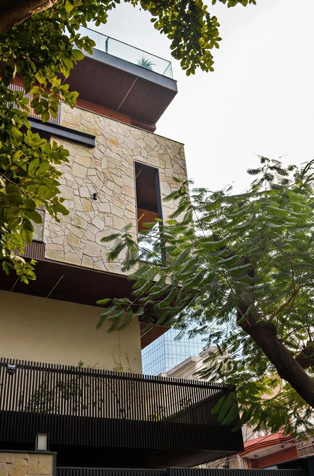 Low-angle view of a modern multi-story building with stone and glass exterior, surrounded by green trees, against a cloudy sky.