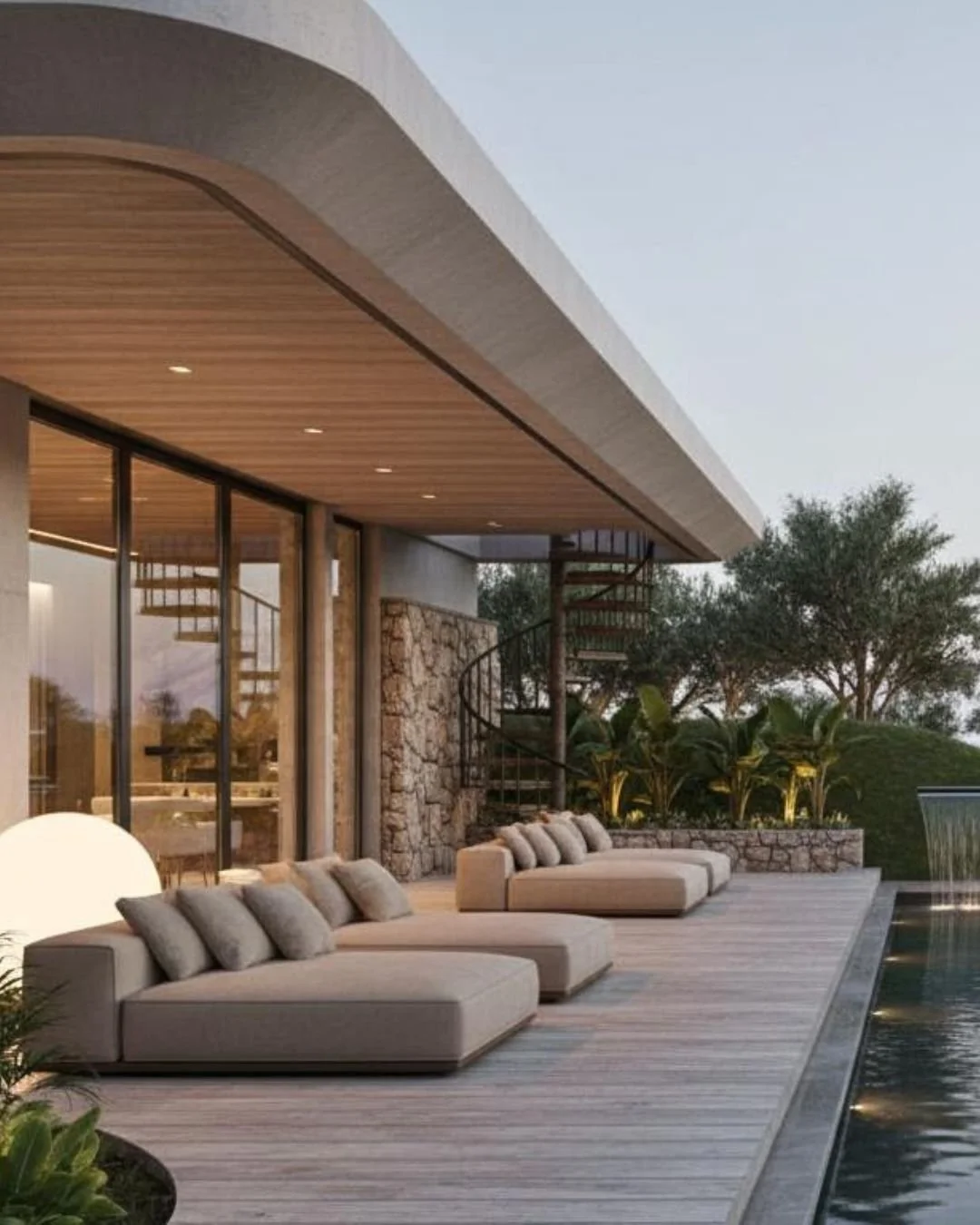 Modern outdoor patio area with beige lounge chairs, potted plants, adjacent to a swimming pool, at sunset, with trees in the background.