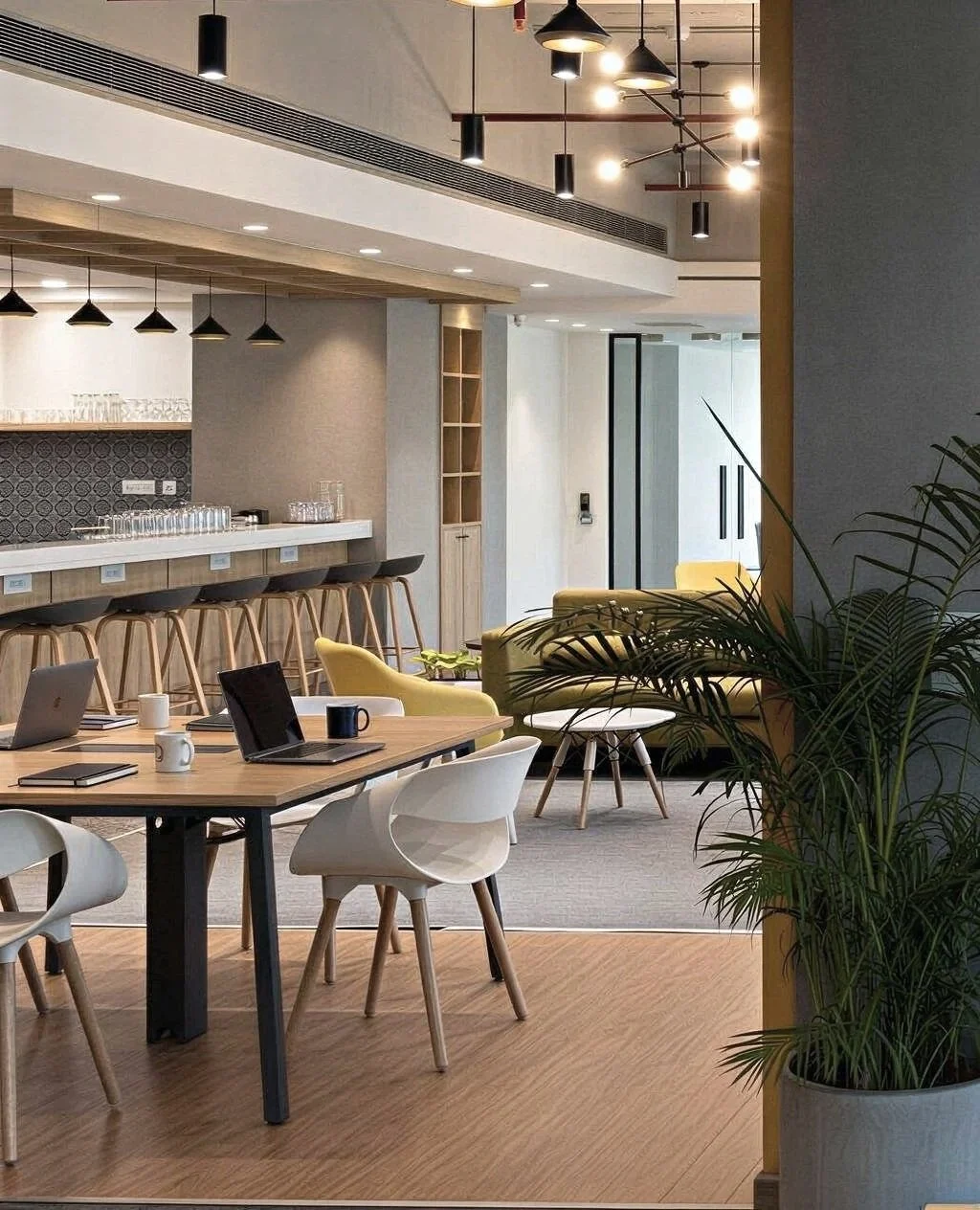 Modern office lounge and meeting area with wooden flooring, yellow and white chairs, and a bar counter with stools, illuminated by pendant and ceiling lights, with potted plants nearby.