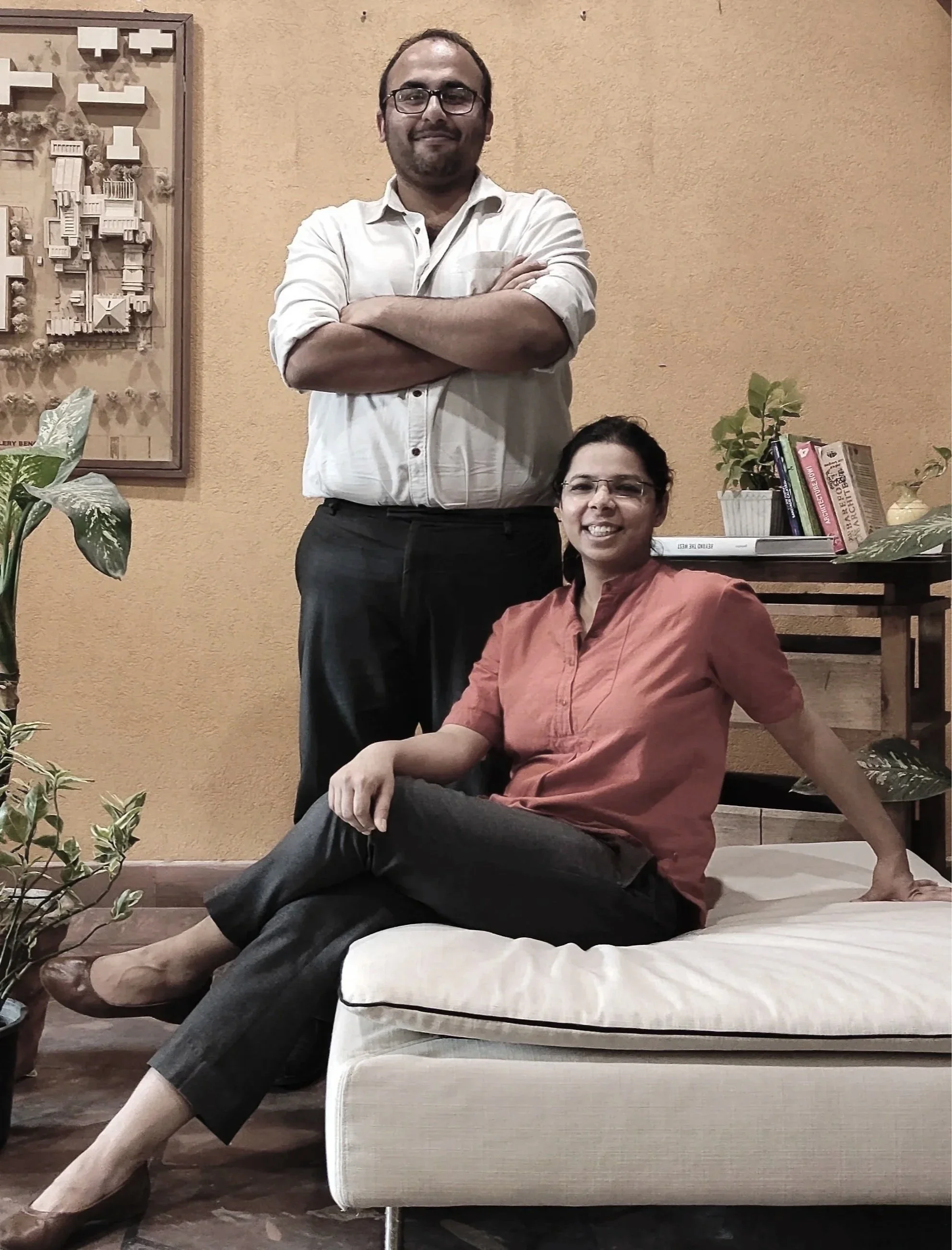 A man with glasses and a beard, wearing a white shirt with rolled-up sleeves, standing with arms crossed, and a woman with glasses, wearing an orange shirt, sitting on a bench indoors, with plants, books, and artwork on the wall behind them.