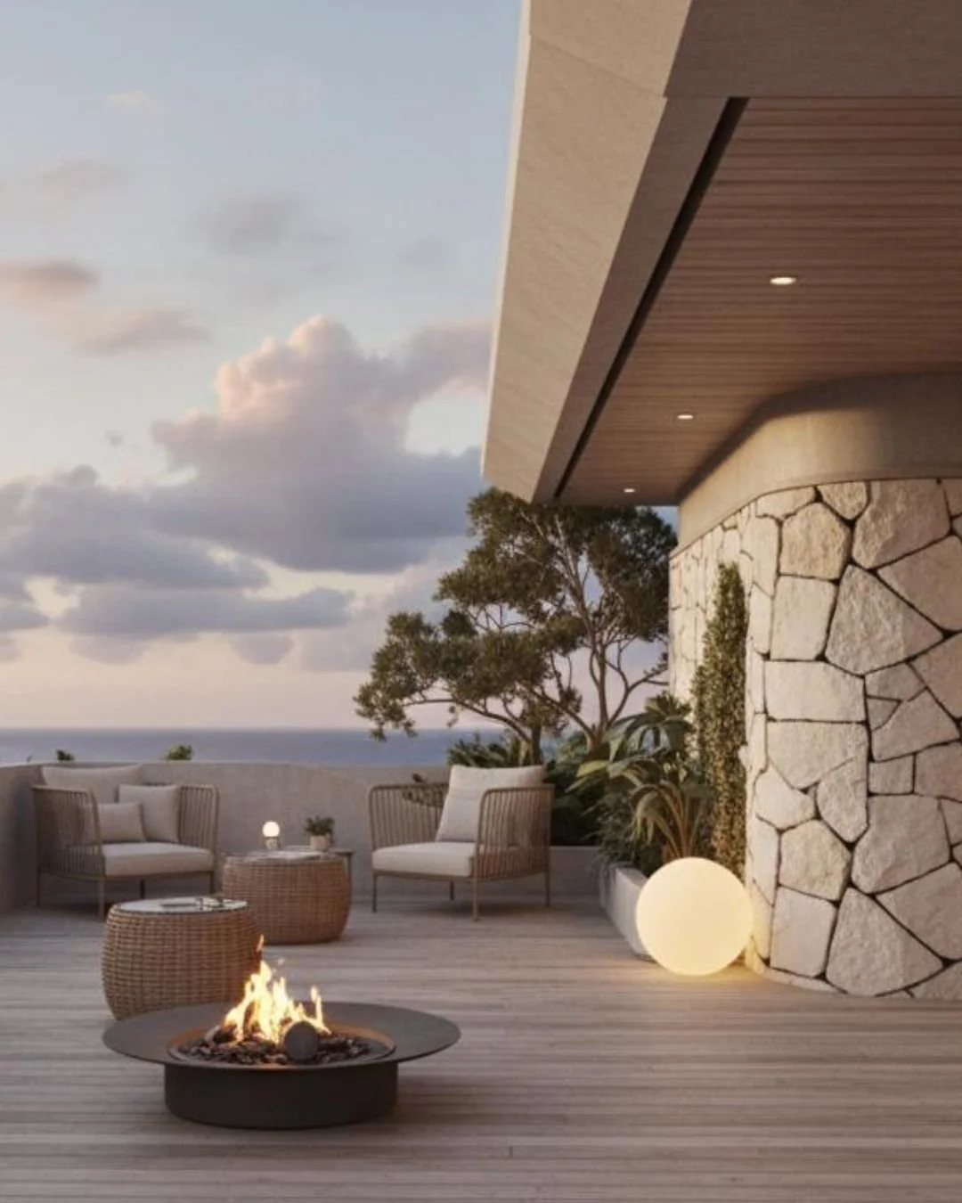 Outdoor patio with modern furniture, a fire pit, round lamps, and trees, overlooking the ocean during sunset.