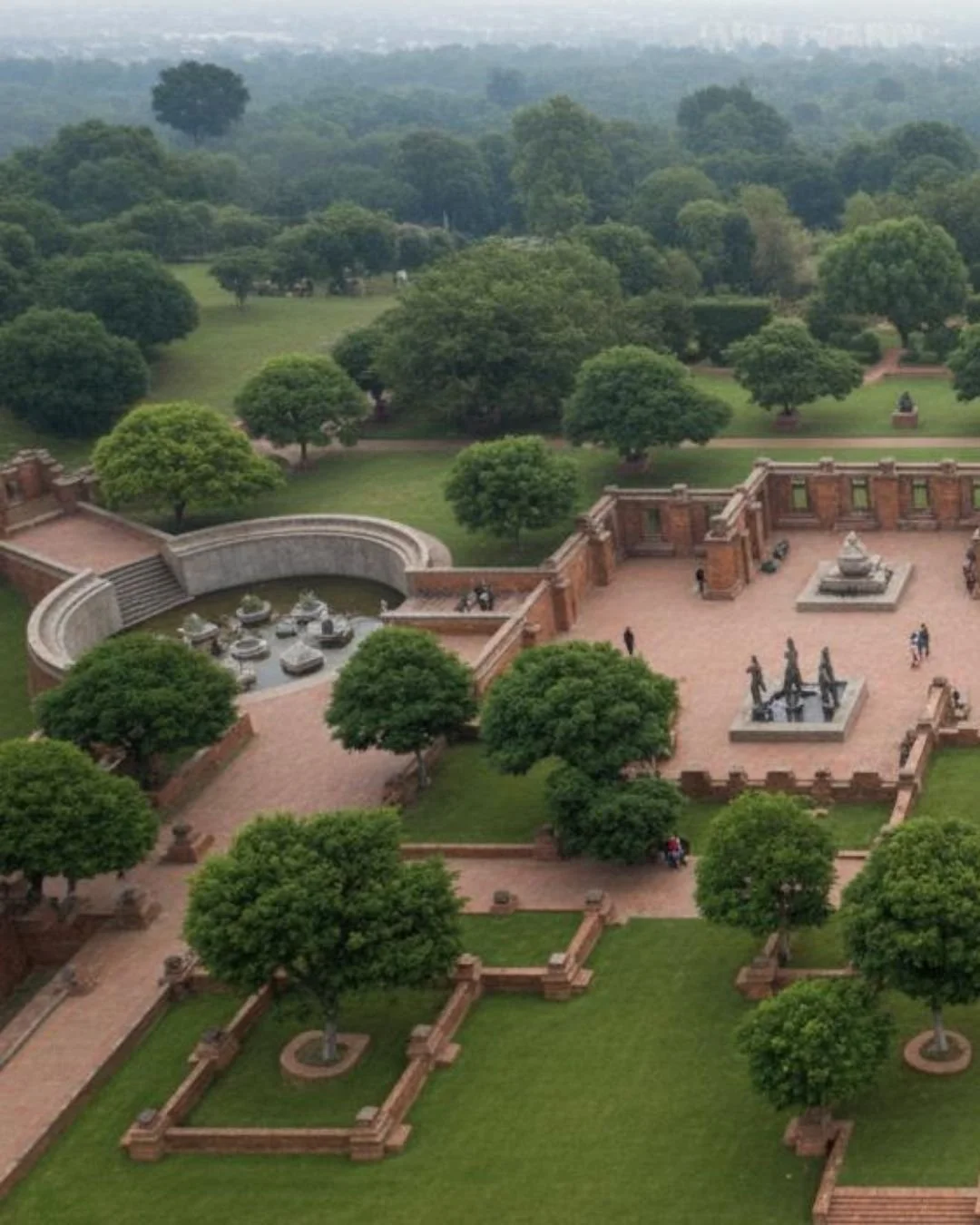 Aerial view of a historic site with a red-brick courtyard, statues, a fountain, and lush green trees, surrounded by a park.