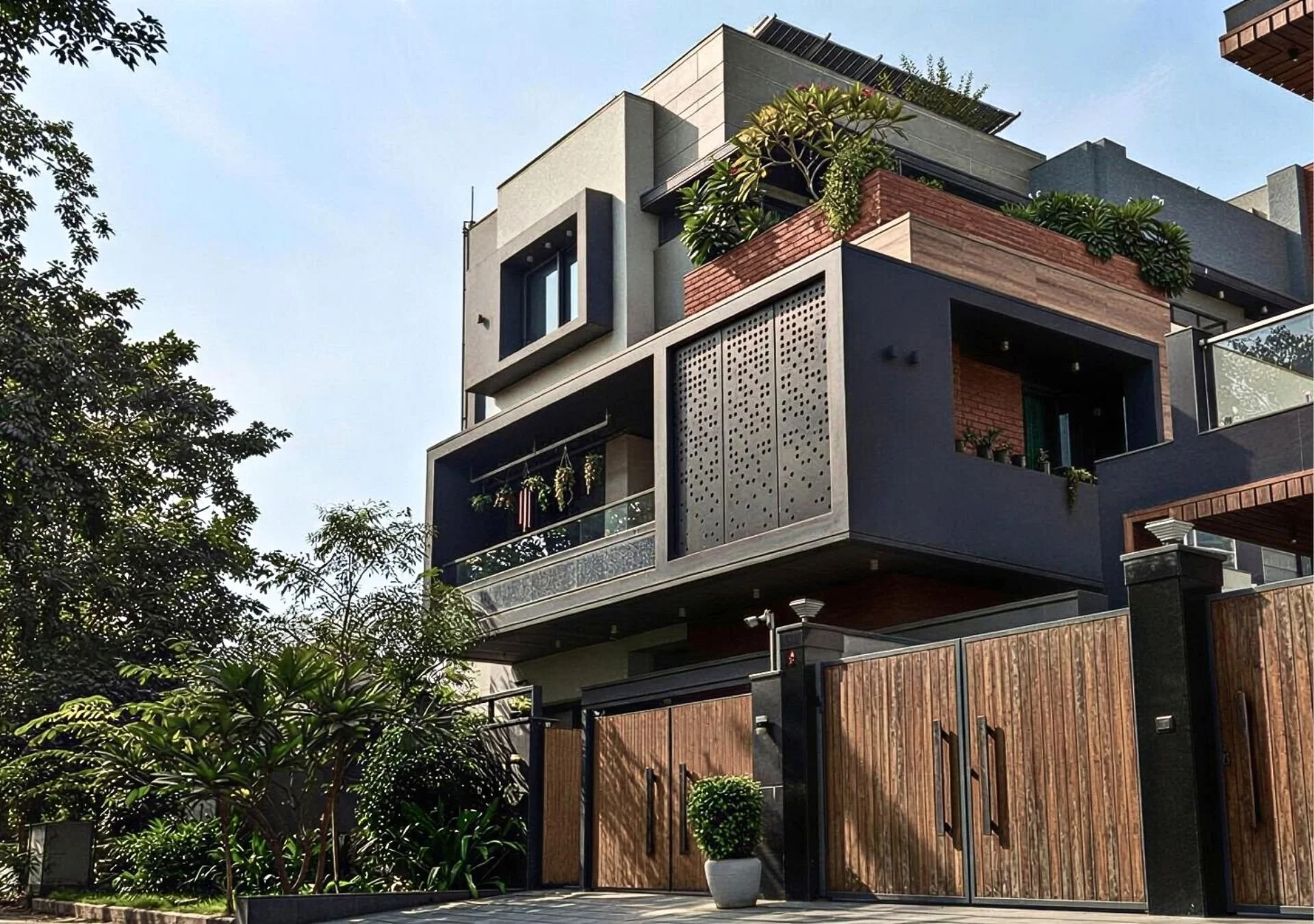 Modern multi-story house with a gated wooden fence, outdoor plants, and spacious balconies, featuring a contemporary architectural style with geometric shapes and large windows, surrounded by lush greenery.