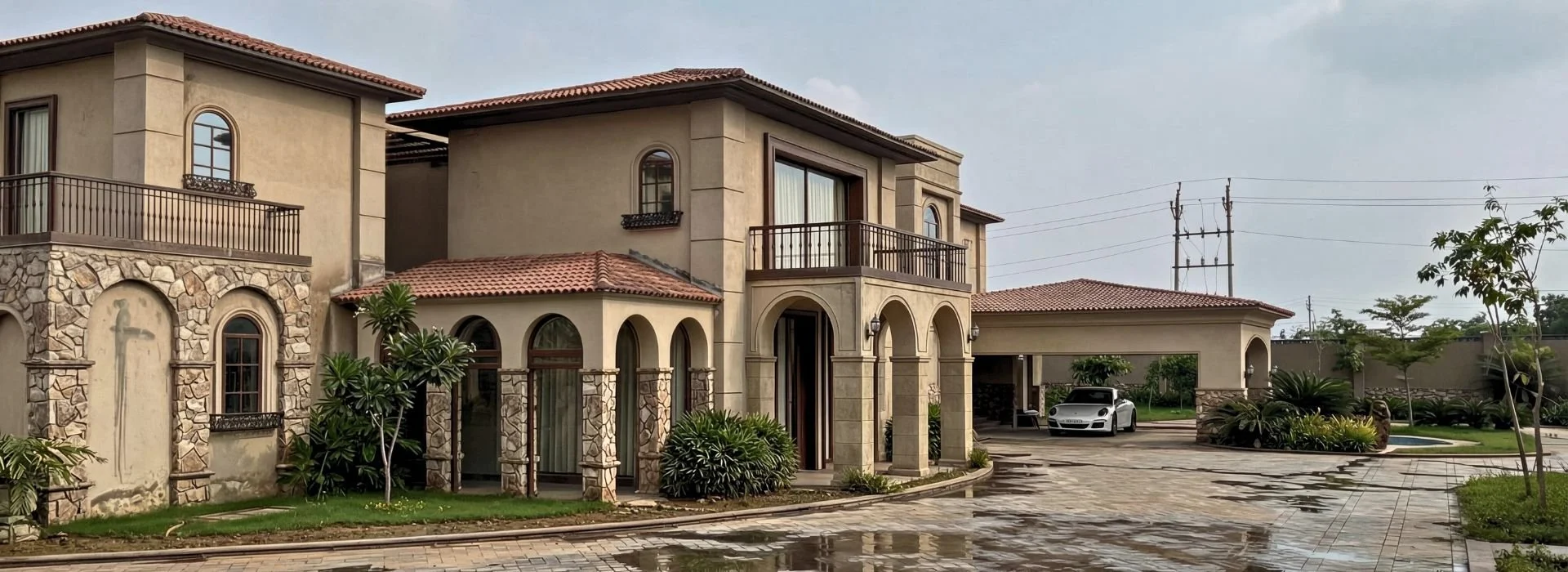 A large, elegant two-story house with arches, stone accents, balconies, a tiled roof, and a driveway with a white luxury car. The front yard has green plants and trees; some puddles suggest recent rain.