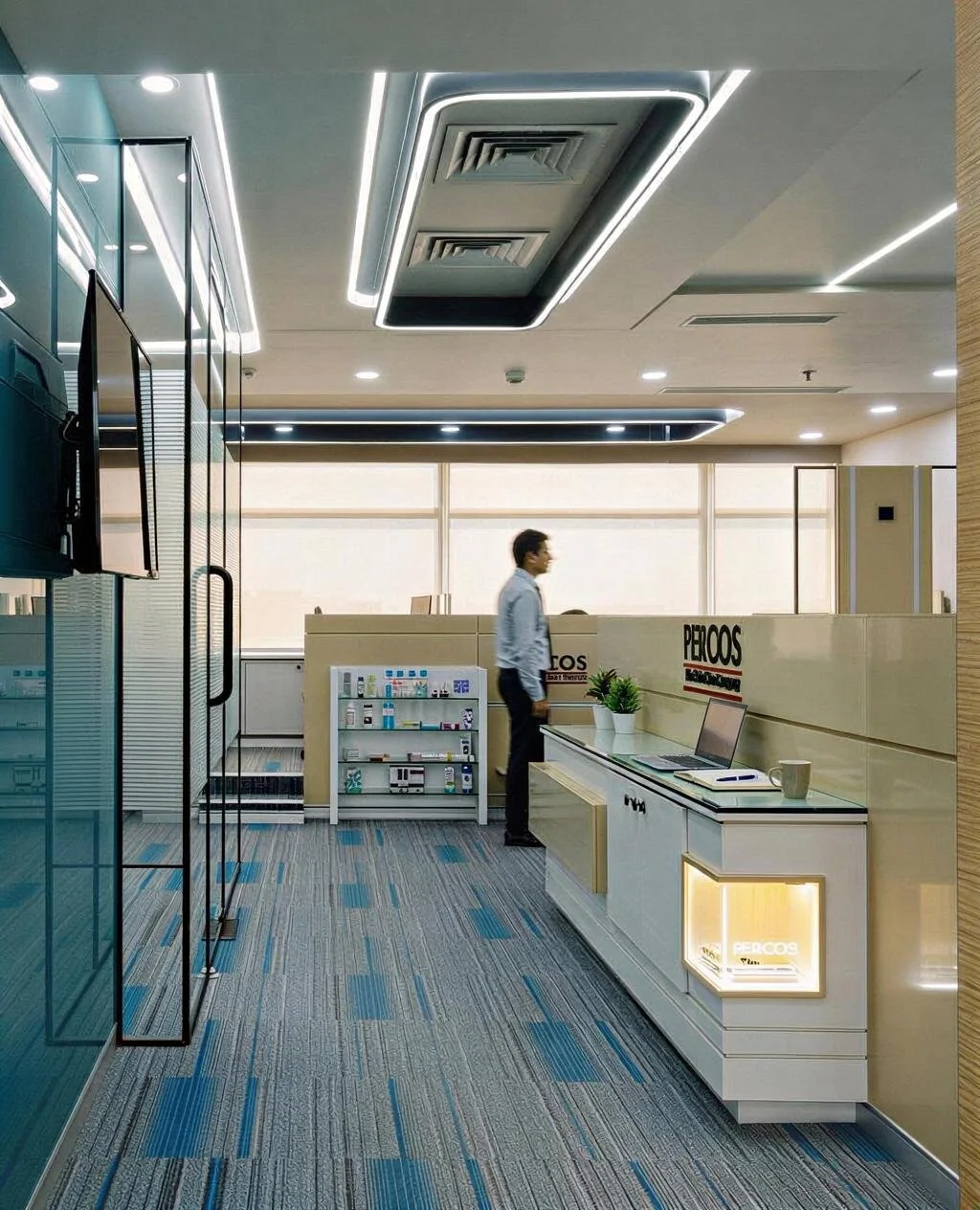 Office reception area with a man walking past a desk, a laptop, a potted plant, and promotional materials. Modern ceiling and large windows letting in natural light.