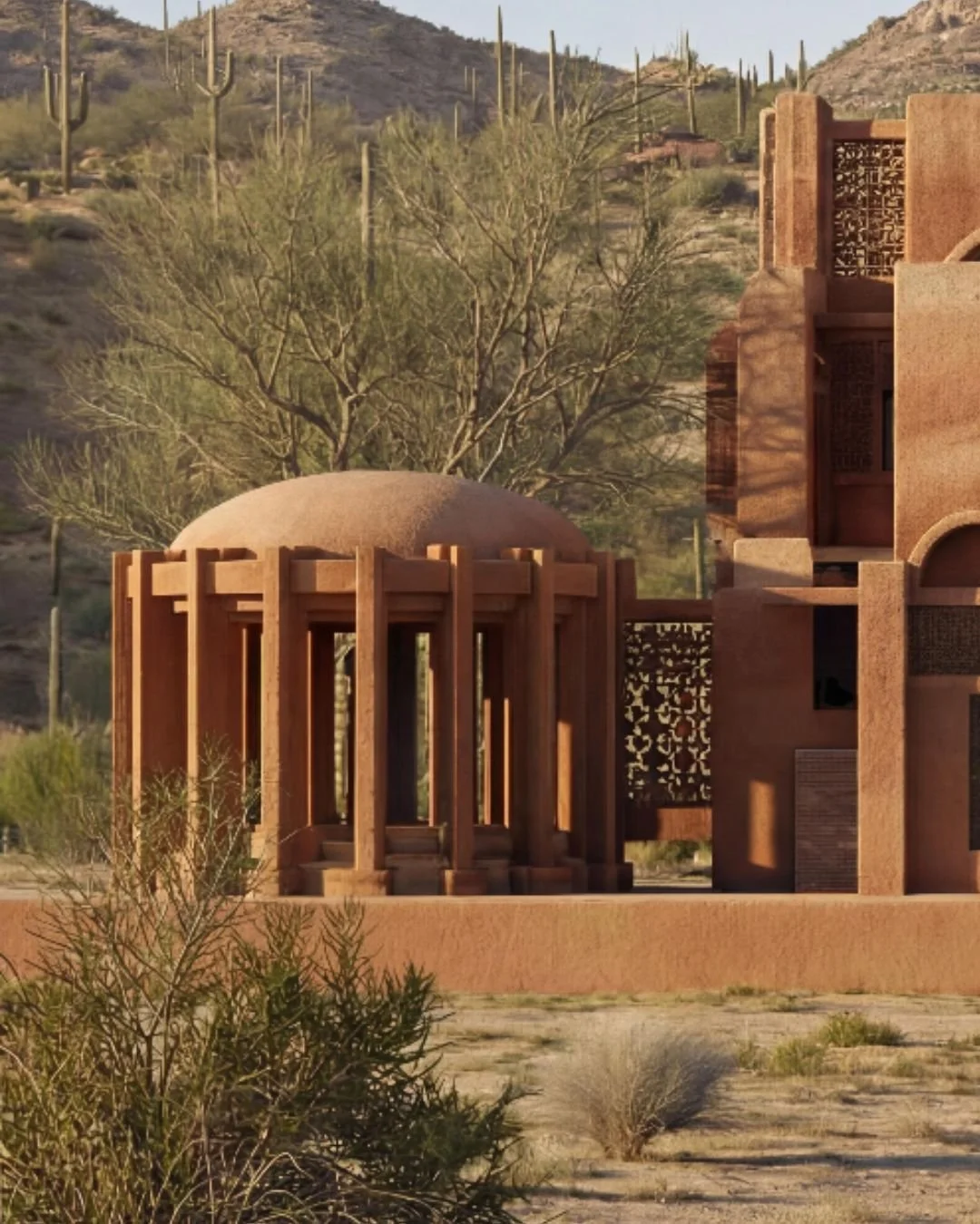 A desert scene with a modern architectural building featuring rounded and angular features, earthy tones, and decorative metalwork. Saguaro cacti and sparse desert vegetation are visible in the background.