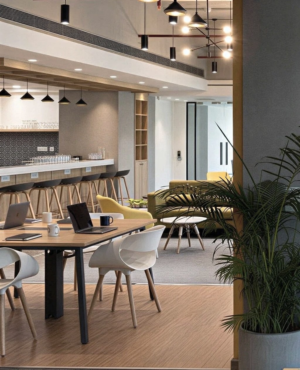 Modern office lounge and meeting area with wooden flooring, yellow and white chairs, and a bar counter with stools, illuminated by pendant and ceiling lights, with potted plants nearby.