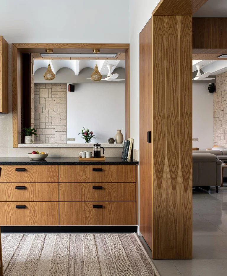 Interior view of a living space with a kitchen area featuring wooden cabinets and countertops, a decorative cutout wall with potted plants and vases, and a glimpse into a living room with a sofa and modern ceiling fans.