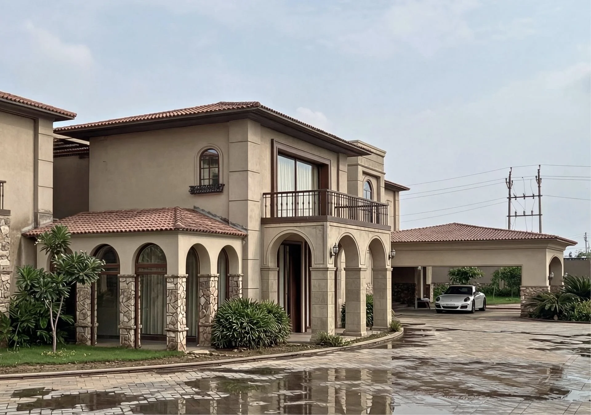 Two-story house with beige walls, red-tile roof, arched windows, and a balcony with a black railing, fenced yard with plants, stone and paved driveway, white luxury car parked under carport, power lines in background.