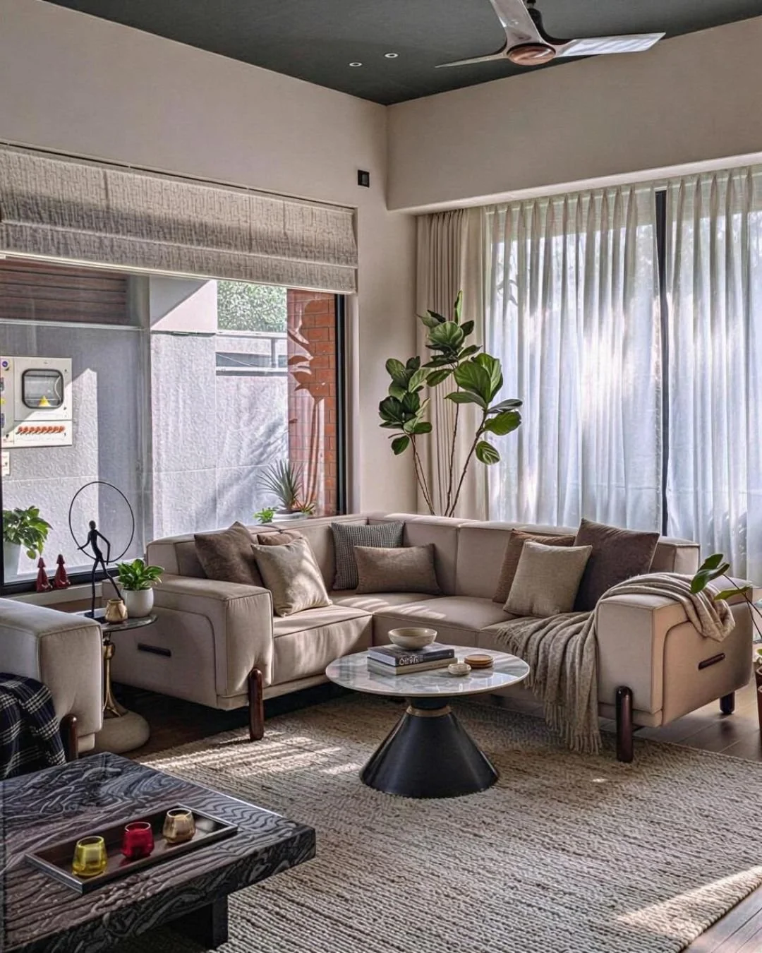 Bright living room with large cream sectional sofa, various pillows, and a round marble coffee table. Large windows with sheer curtains and tall indoor plants add to the cozy atmosphere. Decor includes small sculptures, books, and candles.