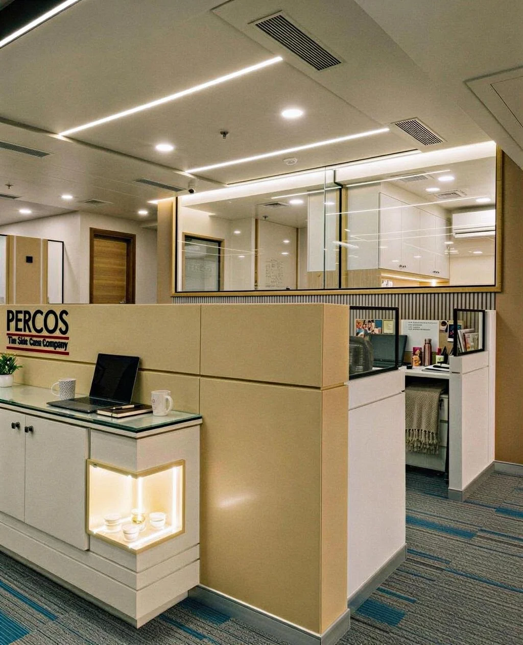 Office reception desk and workspace area with computer, mugs, framed photos, and decorative shelves, featuring a large mirror on the wall and modern lighting.