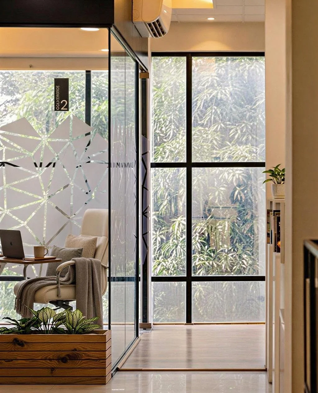 A modern office space with a glass partition, a white office chair with pillows and a blanket, a small table with a laptop and mug, large windows with a view of greenery, and a small potted plant on a shelf.