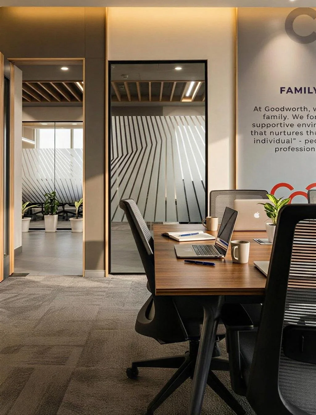 Office conference room with a wooden table, laptops, notebooks, coffee mugs, and potted plants. There are glass walls with stripe patterns and a wall with text about family values.