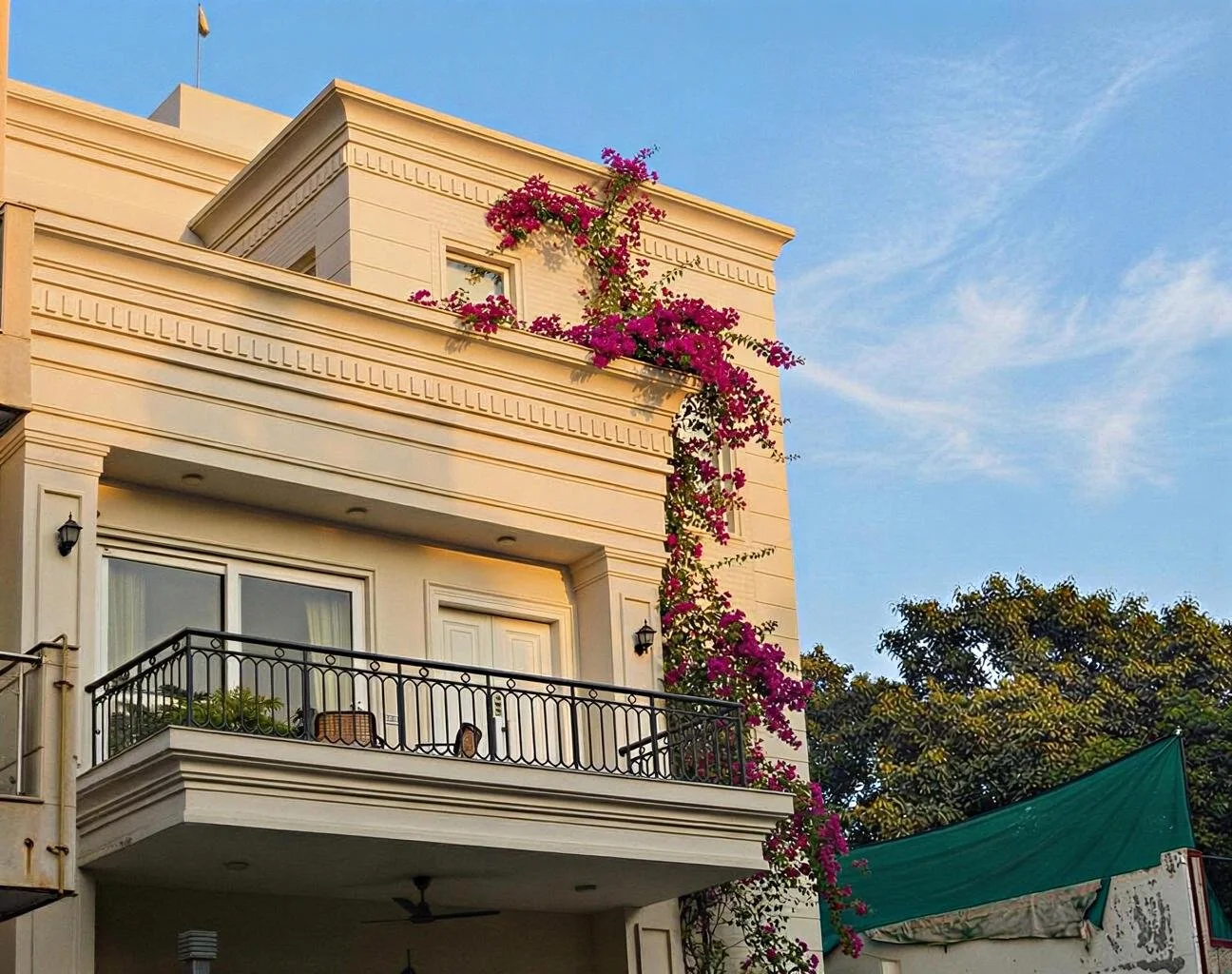 A multi-story beige building with decorative trim, a black wrought-iron balcony on the second floor, and vibrant pink bougainvillea climbing its facade, with a blue sky in the background.