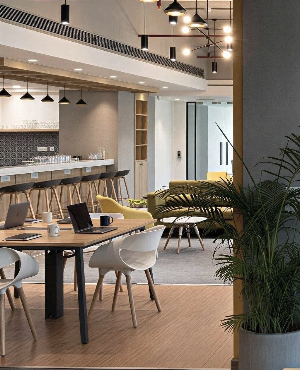 Modern office lounge and meeting area with wooden flooring, yellow and white chairs, and a bar counter with stools, illuminated by pendant and ceiling lights, with potted plants nearby.