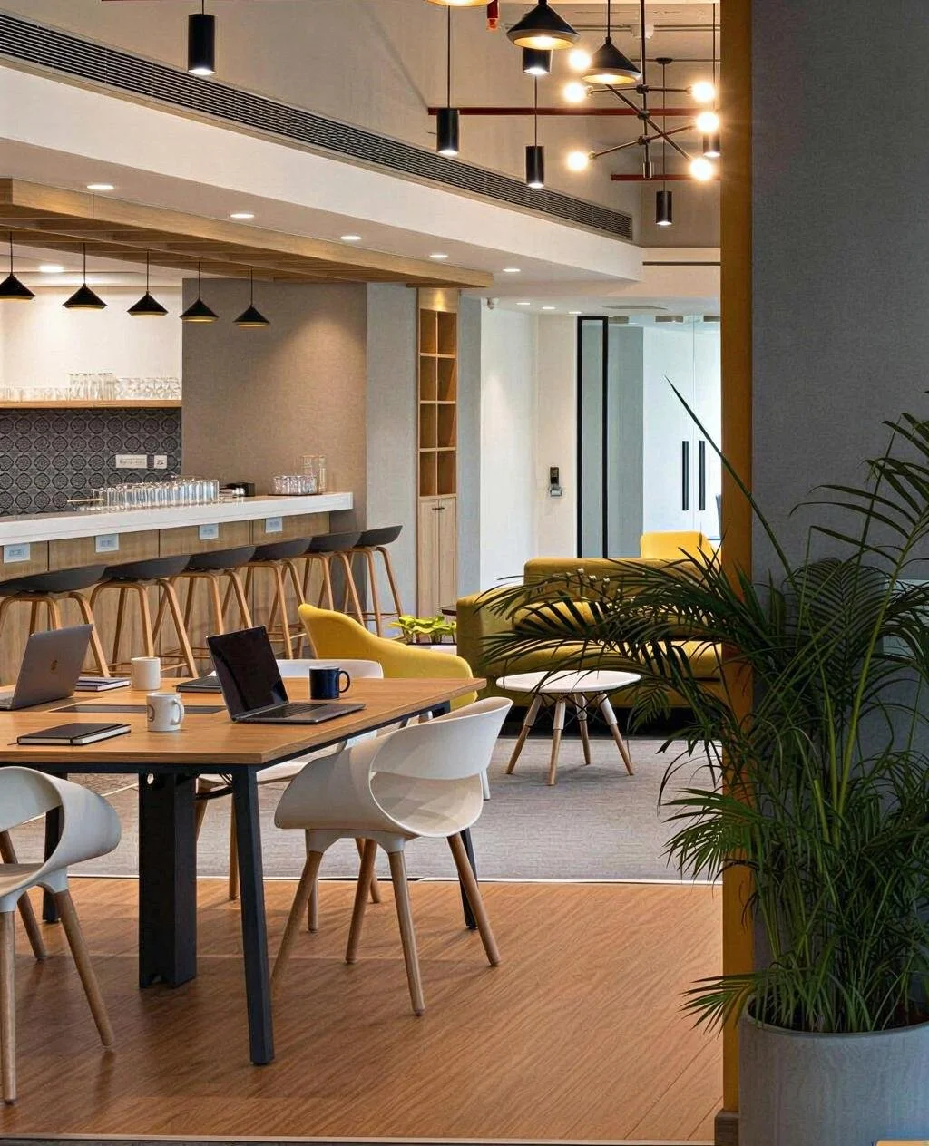 Modern office lounge with a kitchenette, wooden tables, white and yellow seating, laptops, mugs, and potted plants.