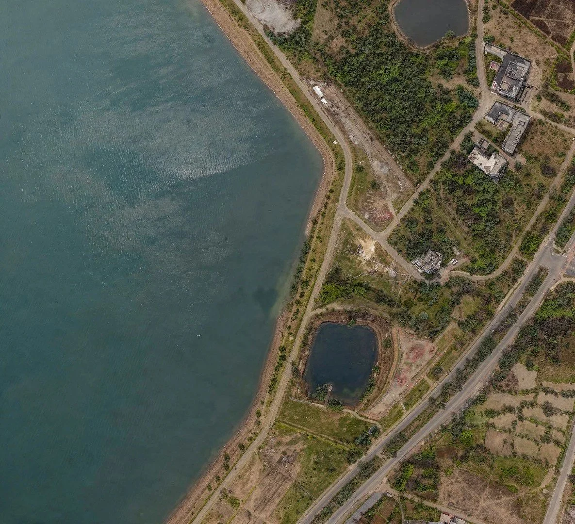 Aerial view of a large body of water next to a shoreline with dirt paths, greenery, and several buildings, with smaller ponds nearby.