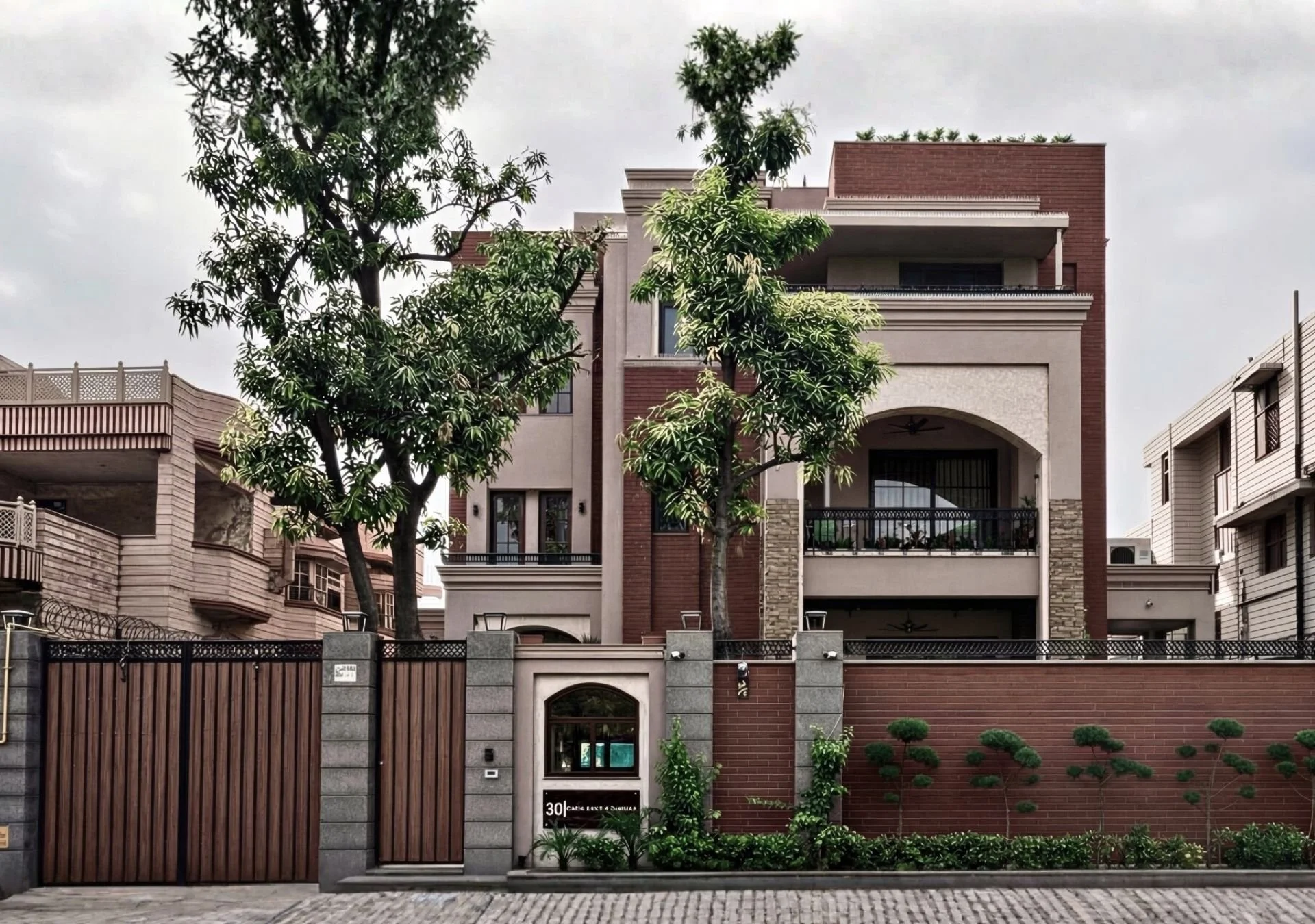 Multi-story modern house with a red brick and beige facade, arched and rectangular windows, balcony with black railings, large trees in front, and a brick and wood fence with greenery along the sidewalk.