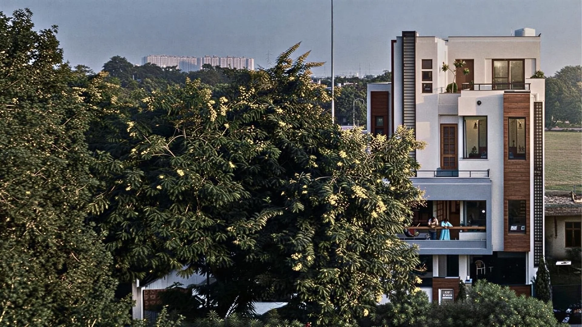 A modern multi-story house with large windows and balconies, partially obscured by lush green trees in the foreground, with a cityscape in the background.