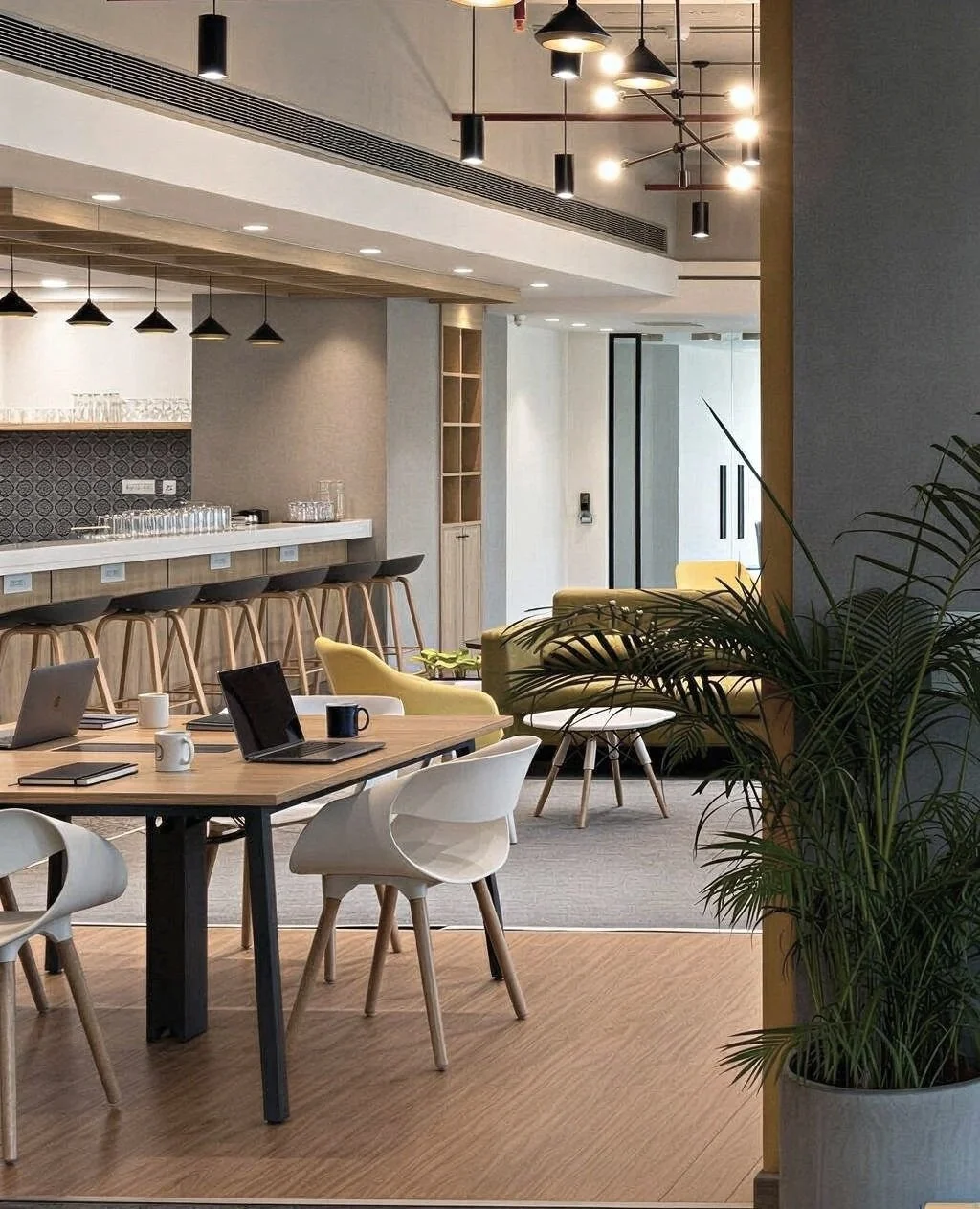 Modern office lounge and meeting area with wooden flooring, yellow and white chairs, and a bar counter with stools, illuminated by pendant and ceiling lights, with potted plants nearby.
