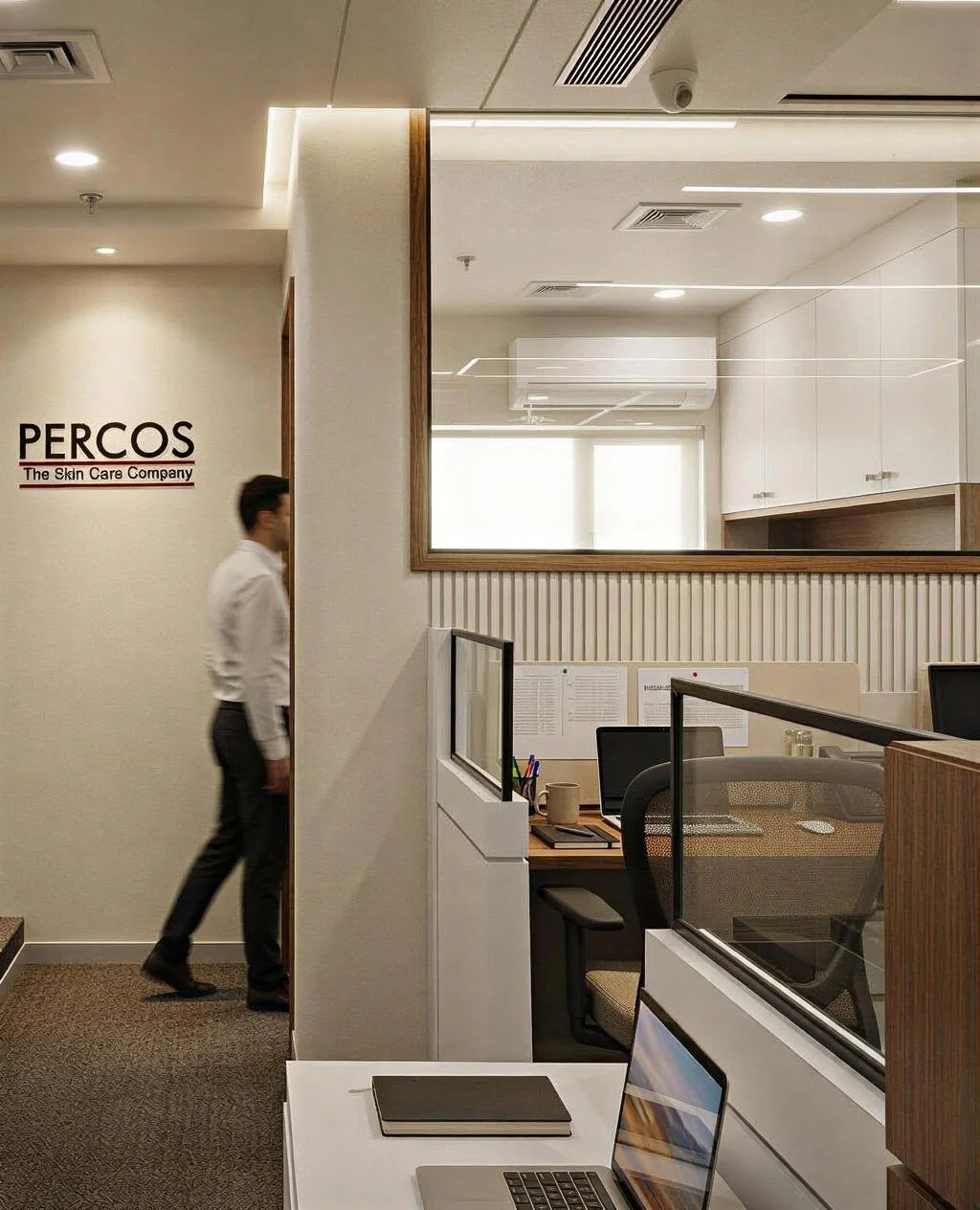 Office reception area with a man walking past, desks with computers and office supplies, and a wall with the sign 'PERCOS The Skin Care Company'.