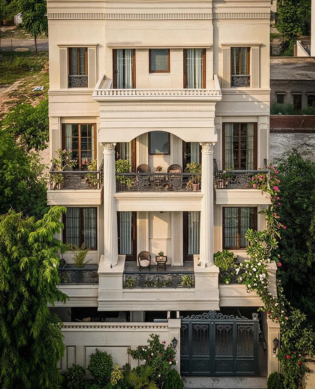 Multi-story beige luxury house with balconies, columns, and ornate railings surrounded by greenery and flowering plants.