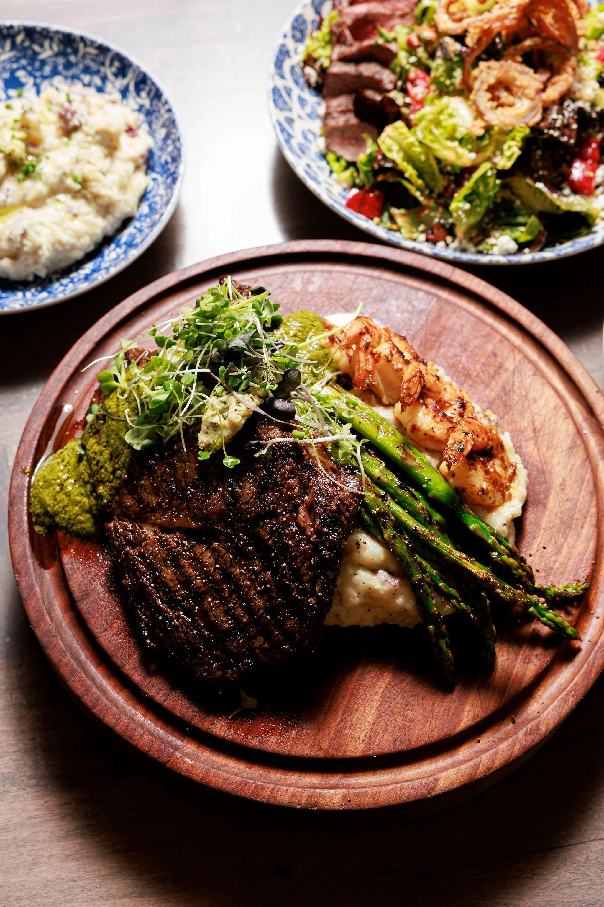 A plate of grilled steak, roasted asparagus, mashed potatoes, and microgreens, served on a wooden platter. In the background, there are two bowls with side dishes, one with potato salad and the other with a mixed salad.