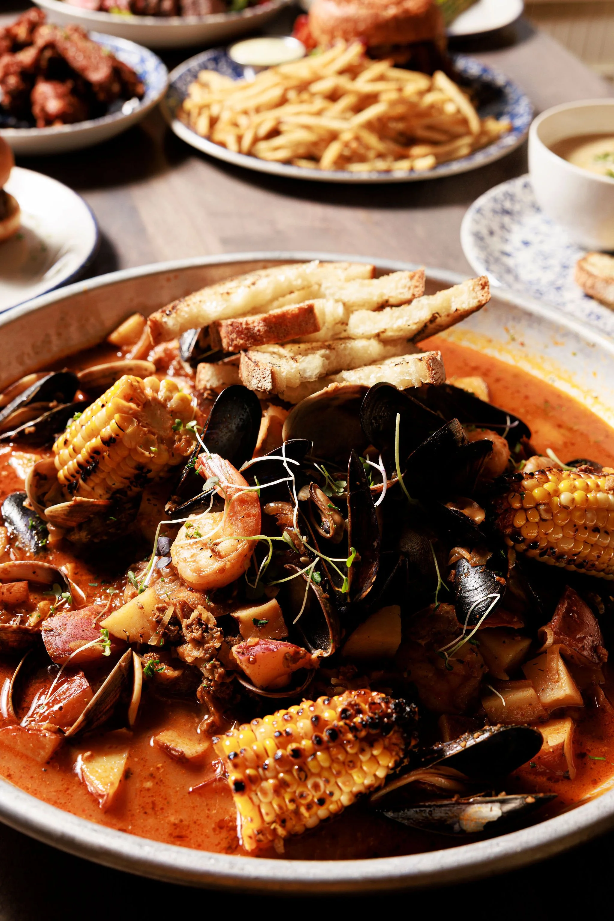 Seafood boil with mussels, corn on the cob, potatoes, and sausage in a spicy broth, garnished with microgreens, served in a shallow bowl.