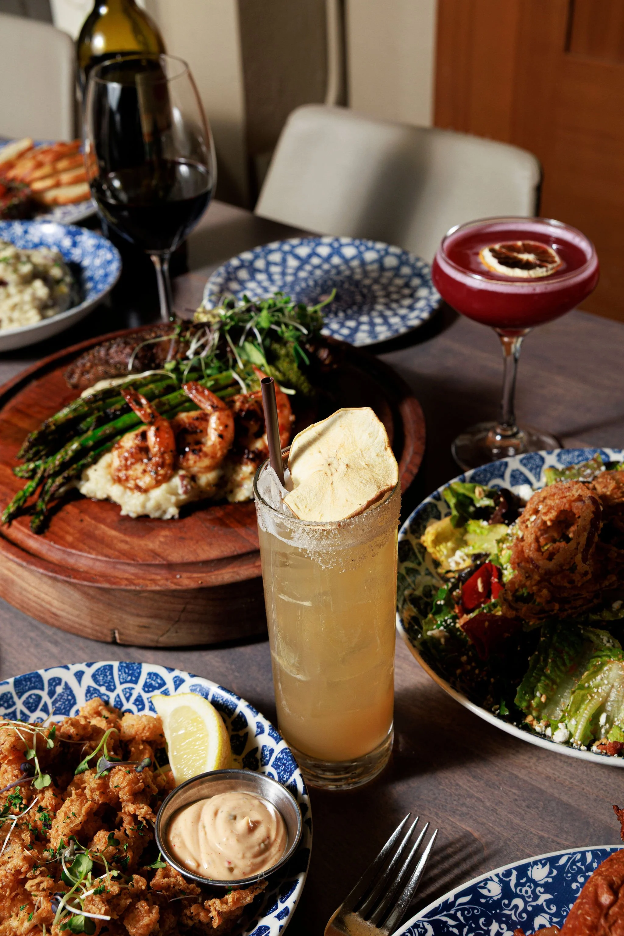 A table set with various dishes including fried chicken, asparagus with a sauce, a seafood dish with lemon, salads, a glass of red wine, a cocktail with a lemon wedge, and a red fruit cocktail with a lemon slicer garnish.