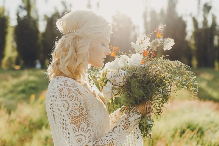  Beautiful Bride close up shot of her outdoors in a field at sunset sniffing the flowers in her wild bouquet. She has short blonde hair in boho curls and natural bridal makeup 