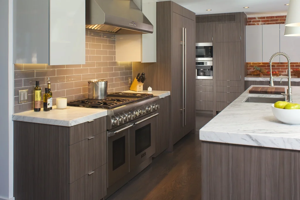 This kitchen was designed with family living, entertaining and cooking in mind. Lots of cabinets and drawers throughout provide convenient space for stowing dishes, pots and pans, table linens and cooking necessities. Integrated appliances include a microwave and upscale coffee maker, and there are appliance panel doors, a hidden television, built in banquette, and a tidy computer station, The high gloss upper cabinets in a soft gray meld beautifully with the wood grain laminate doors and drawers.