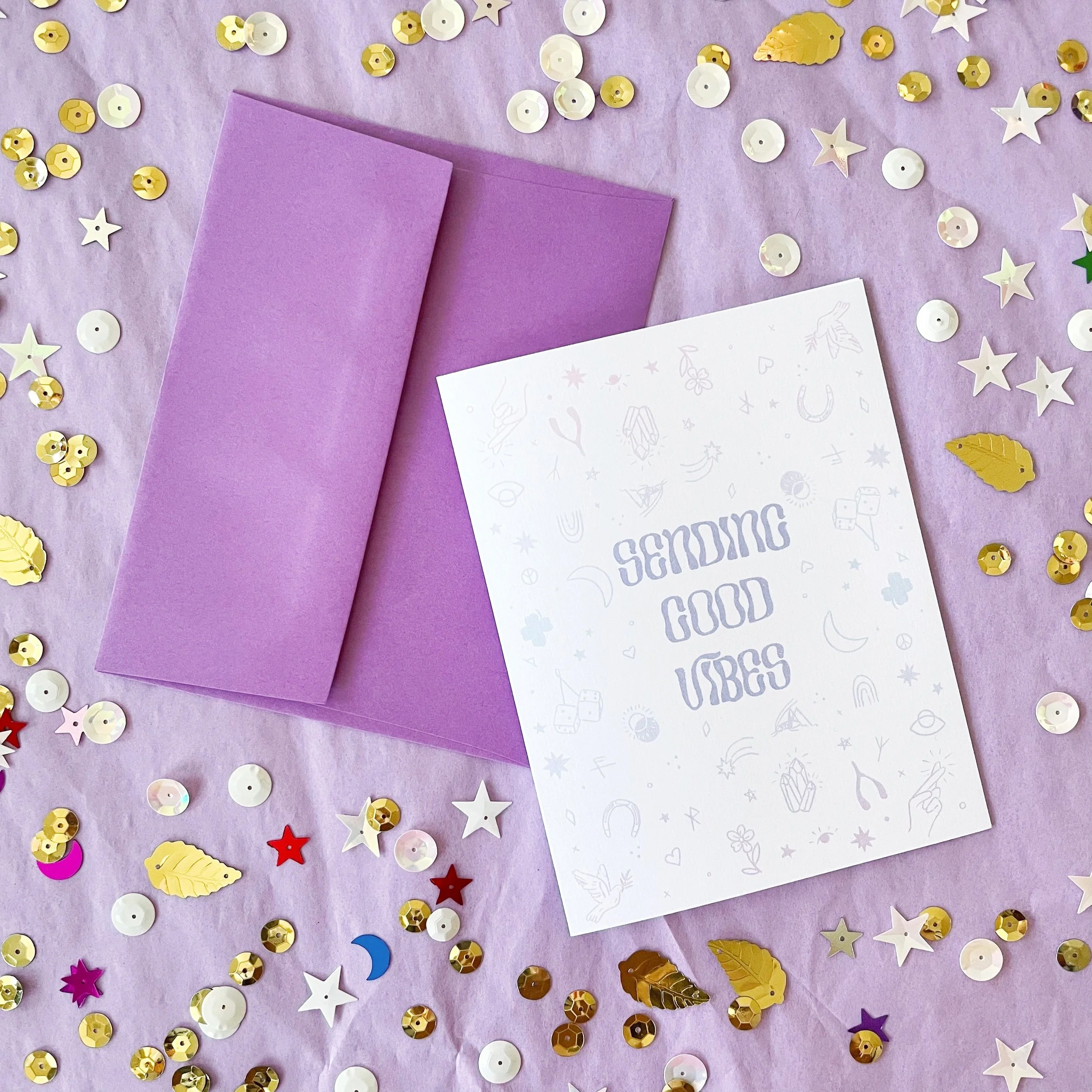 greeting card with drawn good luck charms that says sending good vibes with purple envelope on lavender background with gold and white sequins